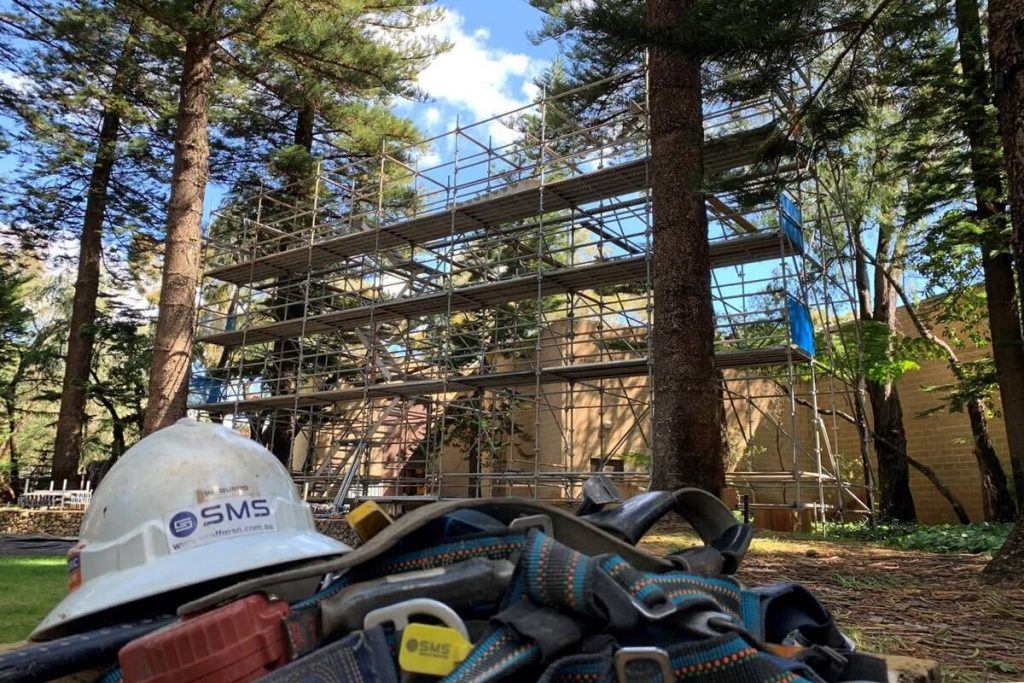 Hard hat and construction gear with SMS company logo, in front of scaffold.