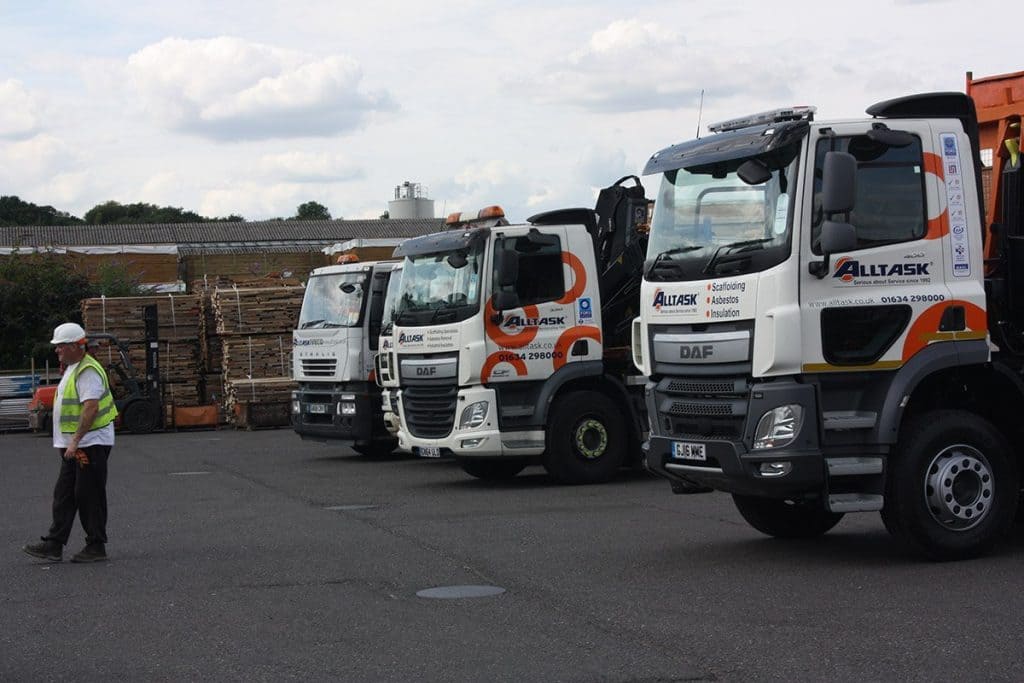 Three Alltask trucks in a row and a man wearing safety gear on a construction site.